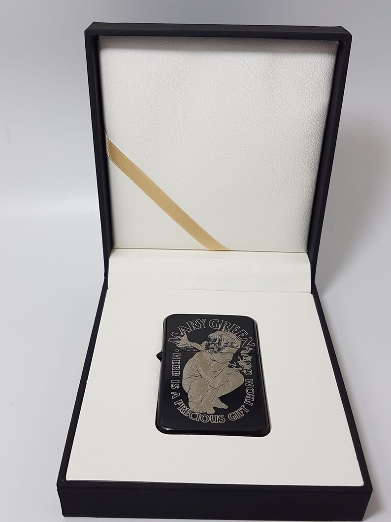 Op de afbeelding: Zwarte aansteker met een zilveren gravure van een vrouw die een plant vasthoudt en de tekst "Mary Green - The Herb is a Precious Gift from God". De aansteker zit in een zwarte doos met een witte binnenkant.
