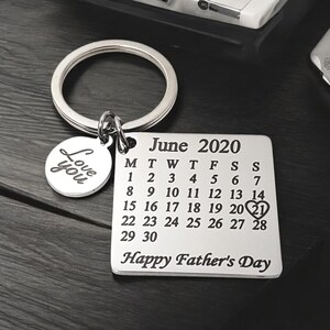 May include: Silver keychain with a calendar charm featuring the month of June 2020 with the 21st circled and a heart. The charm also says "Happy Father's Day". A smaller charm with the words "Love you" is attached to the keychain.