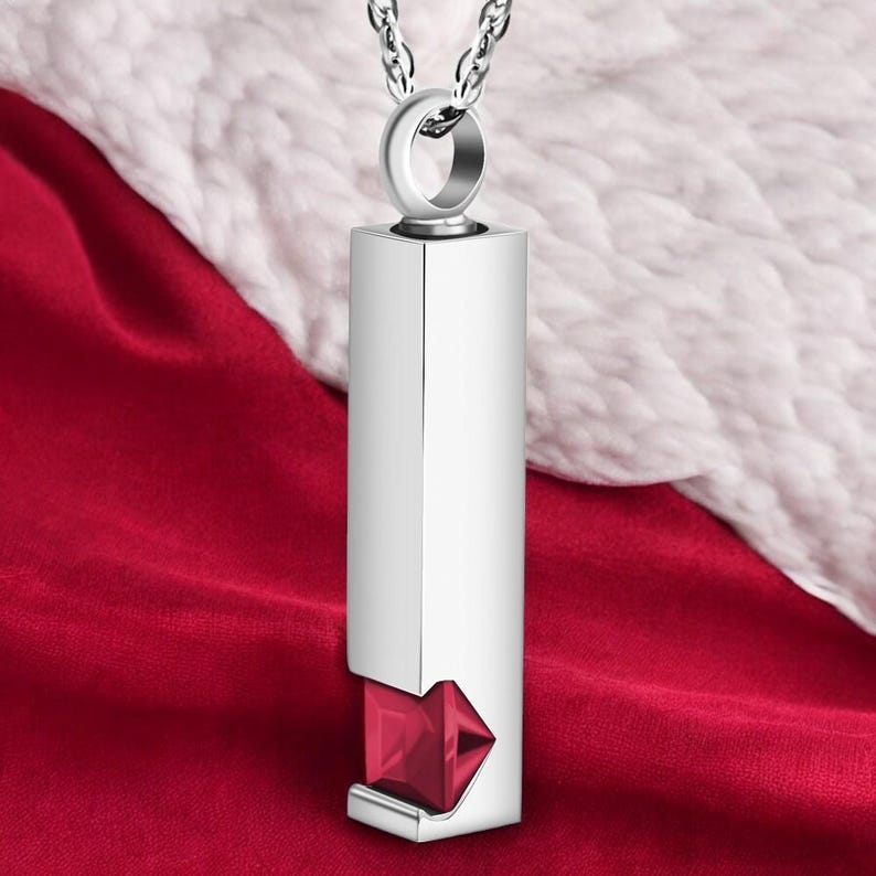 May include: Silver pendant necklace with a red gemstone. The pendant is a rectangular prism with a small opening at the bottom to reveal the gemstone.