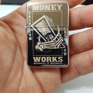 Op de afbeelding: Zilveren aansteker met een zwart-wit afbeelding van een skelet hand die een biljet van &euro; 100 vasthoudt. De aansteker is gegraveerd met de woorden "MONEY WORKS BUSINESS IS BUSINESS".
