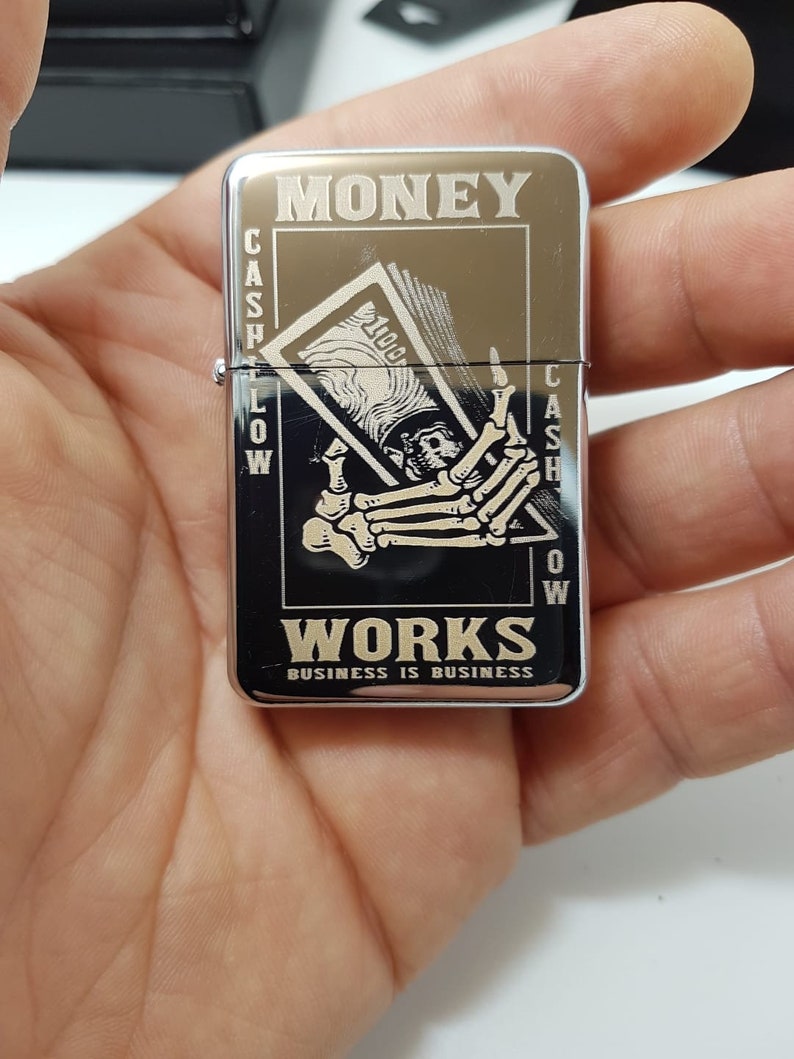 Op de afbeelding: Zilveren aansteker met een zwart-wit afbeelding van een skelet hand die een biljet van &euro; 100 vasthoudt. De aansteker is gegraveerd met de woorden "MONEY WORKS BUSINESS IS BUSINESS".