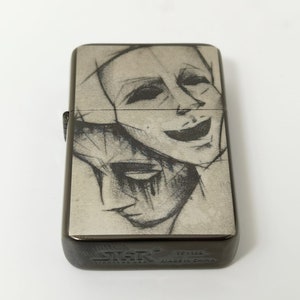 Op de afbeelding: Een zwarte metalen aansteker met een zilveren voorzijde waarop een zwart-witte schets van twee gezichten te zien is. De aansteker heeft de tekst "Zippo" en "Made in China" op de onderkant.