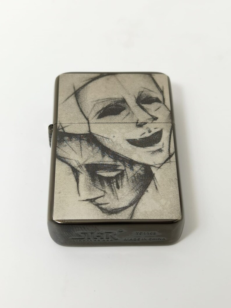 Op de afbeelding: Een zwarte metalen aansteker met een zilveren voorzijde waarop een zwart-witte schets van twee gezichten te zien is. De aansteker heeft de tekst "Zippo" en "Made in China" op de onderkant.