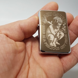 Op de afbeelding: Een zilveren aansteker met een gouden ontwerp met een engel die een marihuanablad vasthoudt. De tekst "Mary Jane" en "God's Gift from the Herb is a Precious Gift" is gegraveerd op de aansteker.