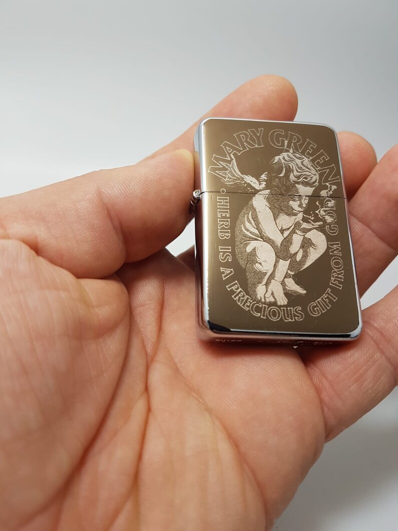 Op de afbeelding: Een zilveren aansteker met een gouden ontwerp met een engel die een marihuanablad vasthoudt. De tekst "Mary Jane" en "God's Gift from the Herb is a Precious Gift" is gegraveerd op de aansteker.