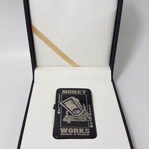 Op de afbeelding: Zwarte aansteker met een zilveren ontwerp van een skelet hand die een stapel geld vasthoudt. De tekst "MONEY WORKS" is onder de afbeelding gedrukt. De tekst "CASHFLOW" is gedrukt aan de linkerkant van de afbeelding en "CASHFLOW" is gedrukt aan de rechterkant van de afbeelding. De tekst "BUSINESS IS BUSINESS" is gedrukt onder de tekst "MONEY WORKS".