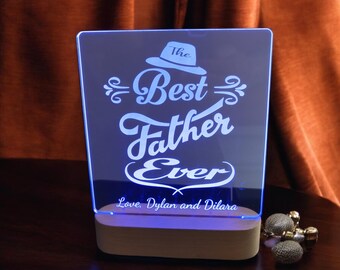May include: A blue LED light with a white wooden base. The light features a white inscription that reads "The Best Father Ever" with a hat graphic above the text. The inscription also includes "Love, Dylan and Dilara" at the bottom.