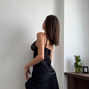 Puede incluir: Una mujer con un vestido de sat&eacute;n negro con ribete de encaje. Est&aacute; de pie frente a una pared blanca y mirando hacia arriba.