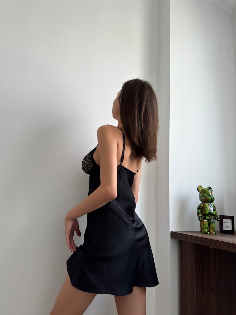 Puede incluir: Una mujer con un vestido de sat&eacute;n negro con ribete de encaje. Est&aacute; de pie frente a una pared blanca y mirando hacia arriba.