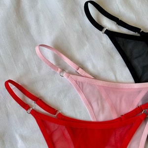 May include: Three pairs of sheer mesh underwear with adjustable straps. The underwear is in red, pink, and black.