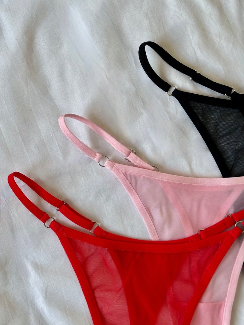 May include: Three pairs of sheer mesh underwear with adjustable straps. The underwear is in red, pink, and black.