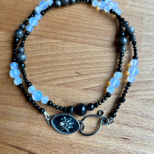 Puede incluir: Un collar de cuentas negras y blancas con un dije de araña de plata. El collar está hecho con cuentas de ónix negro, ópalo blanco y hematites gris.