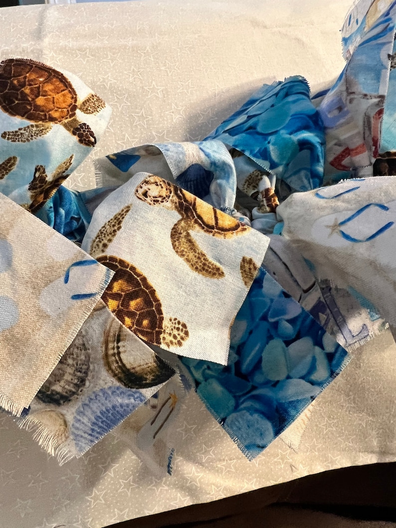 May include: Close-up of fabric squares with a coastal theme. The squares feature sea turtles, seashells, and blue water patterns. The fabric has a light blue and white background with a star pattern.