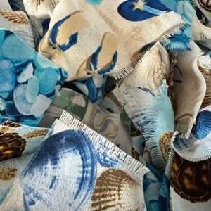 May include: Close-up of fabric scraps with a beach theme. The fabric features seashells, starfish, and turtles in shades of blue, tan, and white. The edges of the fabric are frayed, giving a textured appearance. The overall theme is coastal.