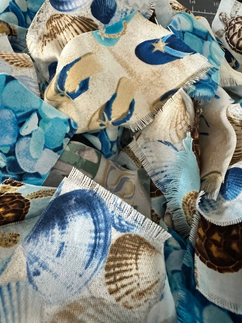 May include: Close-up of fabric scraps with a beach theme. The fabric features seashells, starfish, and turtles in shades of blue, tan, and white. The edges of the fabric are frayed, giving a textured appearance. The overall theme is coastal.
