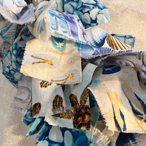 May include: Close-up of fabric scraps with various sea-themed patterns. The fabrics feature images of sea turtles, seashells, starfish, and blue pebble designs. The color palette includes shades of blue, brown, and beige, creating a coastal aesthetic.