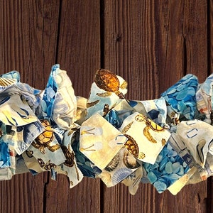 May include: A decorative garland made of fabric strips in shades of blue, white, and tan. The fabric features patterns of turtles, seashells, and other nautical elements. The garland is arranged in a wavy pattern against a brown wooden background.
