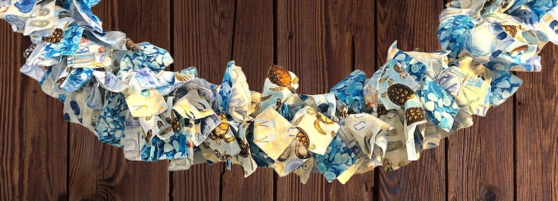 May include: A decorative garland made of fabric strips in shades of blue, white, and tan. The fabric features patterns of turtles, seashells, and other nautical elements. The garland is arranged in a wavy pattern against a brown wooden background.