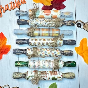 May include: A set of seven wooden rolling pins with various fall-themed designs, including pumpkins, leaves, and plaid patterns. The rolling pins are tied with twine and arranged vertically on a white wooden surface.