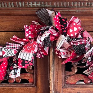 May include: A Valentine's Day garland featuring a mix of red, black, and white ribbons with heart patterns and plaid designs. The garland is draped across a wooden surface, creating a festive and decorative display.
