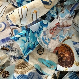 May include: Close-up of fabric scraps with a beach theme. The fabric pieces feature various patterns, including flip-flops, seashells, and turtles, in shades of blue, tan, and white. The edges of the fabric are frayed, adding texture.