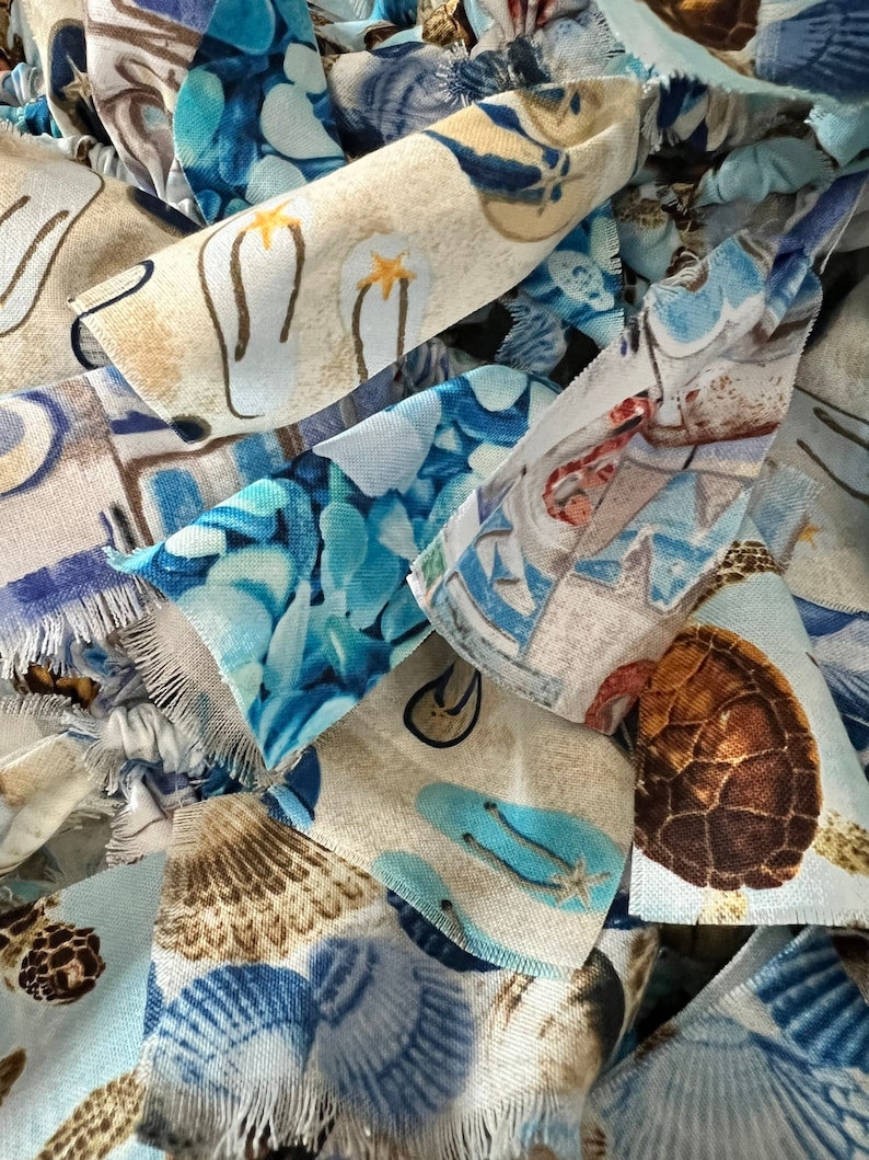 May include: Close-up of fabric scraps with a beach theme. The fabric pieces feature various patterns, including flip-flops, seashells, and turtles, in shades of blue, tan, and white. The edges of the fabric are frayed, adding texture.