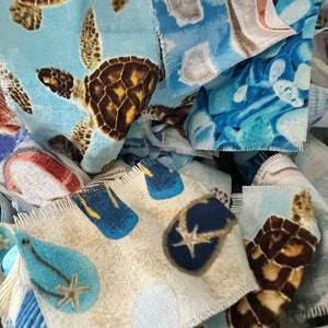 May include: Close-up of fabric scraps with beach-themed patterns. The designs include sea turtles, flip-flops, and starfish in shades of blue, brown, and beige. The edges of the fabric are frayed.