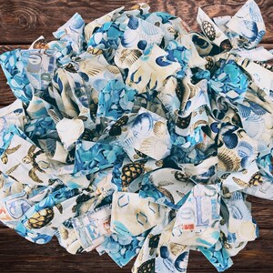 May include: A circular arrangement of fabric strips in shades of blue, white, and tan, featuring seashell and turtle patterns. The fabric is layered and textured, creating a visually appealing, beach-themed design. A few seashells are interspersed within the fabric.