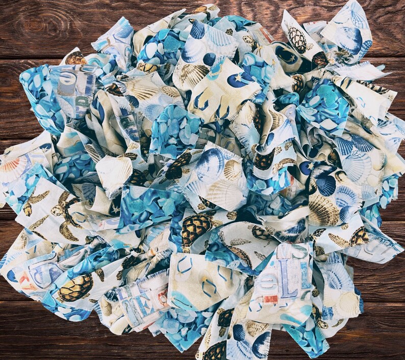 May include: A circular arrangement of fabric strips in shades of blue, white, and tan, featuring seashell and turtle patterns. The fabric is layered and textured, creating a visually appealing, beach-themed design. A few seashells are interspersed within the fabric.