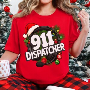 May include: A red short-sleeved t-shirt featuring a graphic of black headphones wearing a Santa hat, surrounded by pine branches and Christmas ornaments. The text 911 DISPATCHER is printed in white across the center of the headphones.