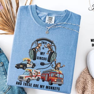 May include: Light blue washed denim t-shirt with a graphic featuring monkeys, headphones, and emergency vehicles. Text reads "THIS IS MY CIRCUS AND THESE ARE MY MONKEYS." The shirt has a crew neck and a Comfort Colors tag.