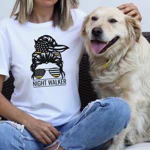 May include: White t-shirt featuring a black and yellow graphic of a woman with a bun, sunglasses, and a bandana. The text "NIGHT WALKER" is below the graphic.
