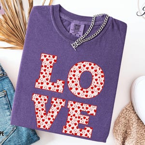May include: A purple Comfort Colors t-shirt with the word "LOVE" in large, block letters. The letters are filled with a pattern of red hearts and headphones. A silver chain necklace is draped across the shirt.