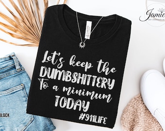 May include: A black t-shirt featuring white text that reads Let's keep the DUMBSHITTERY To a minimum TODAY and #911LIFE. The shirt has a round neckline and a silver sunburst pendant necklace resting on the fabric near the collar area.