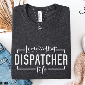 May include: Dark grey heather t-shirt with the text "Livin' that Dispatcher Life" in white. A silver necklace with a sunburst pendant is on the shirt. The shirt is folded with a pair of blue jeans and white sneakers in the background.