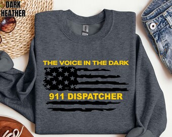 May include: A dark heather gray crewneck sweatshirt featuring a graphic of a distressed American flag in black. The text in yellow reads THE VOICE IN THE DARK above the flag and 911 DISPATCHER below the flag. The garment has long sleeves.