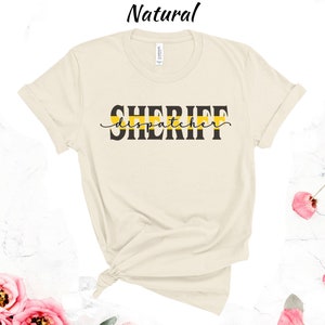 May include: A beige t-shirt with a black graphic that says "SHERIFF dispatcher" with a yellow stripe above the text.