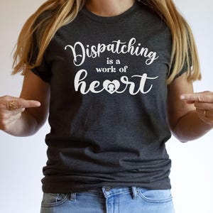 May include: A dark gray t-shirt with the text "Dispatching is a work of heart" in white lettering. A small white heart with a headset inside is included in the text.