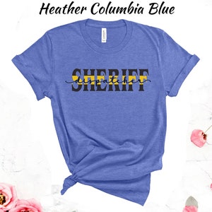May include: A heather Columbia blue t-shirt with the text "SHERIFF dispatcher" in black lettering with a yellow stripe across the word "SHERIFF".