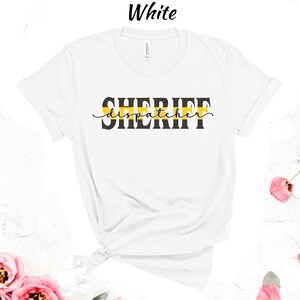 May include: White t-shirt with black text that reads "SHERIFF dispatcher" with a yellow stripe above the text.