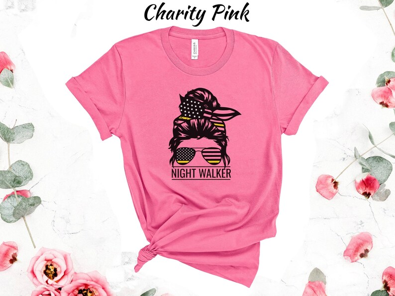 May include: Pink t-shirt featuring a black and white graphic of a woman's hair in a bun with sunglasses and a bandana. The bandana has the American flag design. The text "NIGHT WALKER" is below the graphic.
