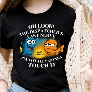 May include: Black t-shirt with a humorous graphic featuring two cartoon fish. One is blue and smiling, the other is orange and wearing a headset, with the text "OH LOOK! THE DISPATCHER'S LAST NERVE I'M TOTALLY GONNA TOUCH IT."