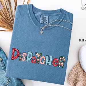 May include: Blue jean colored t-shirt with the word "DISPATCHER" embroidered in red, pink, and green. A silver necklace with a circular pendant rests on the shirt. The shirt is folded on top of a pair of blue jeans.