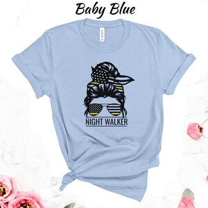 May include: A light blue t-shirt featuring a graphic of a woman's hair in a bun with a bandana and sunglasses. The bandana and sunglasses are decorated with the American flag. The text "NIGHT WALKER" is below the graphic.