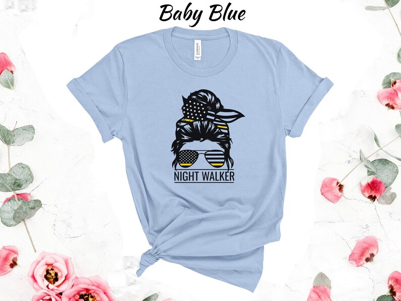 May include: A light blue t-shirt featuring a graphic of a woman's hair in a bun with a bandana and sunglasses. The bandana and sunglasses are decorated with the American flag. The text "NIGHT WALKER" is below the graphic.