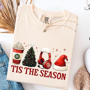 May include: Ivory-colored t-shirt with a festive graphic. The design includes a coffee cup, Christmas tree, headphones, and Santa hat, with the text "TIS THE SEASON" in red. The shirt is made of a soft material.