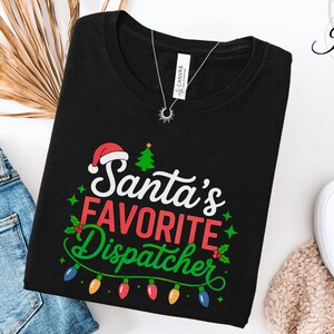May include: Black t-shirt with the text "Santa's Favorite Dispatcher" in white, red, and green. The design includes a Santa hat, Christmas tree, and string of lights. The shirt is folded on a surface with blue jeans and white shoes.
