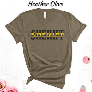 May include: A heather olive t-shirt with the text "SHERIFF dispatcher" in black lettering with a yellow stripe across the text.