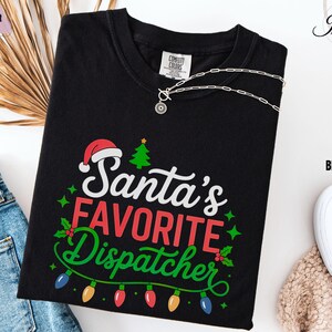 May include: Black t-shirt with the text "Santa's Favorite Dispatcher" in white, red, and green, with a Santa hat and Christmas tree graphic. The shirt has a crew neck and is folded. A silver necklace is draped across the shirt.