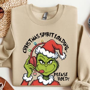 May include: Beige sweatshirt with a Grinch graphic wearing a Santa hat and headset. The text reads "Christmas Spirit Loading... Please Hold!" The Grinch is green with a red and white Santa hat.
